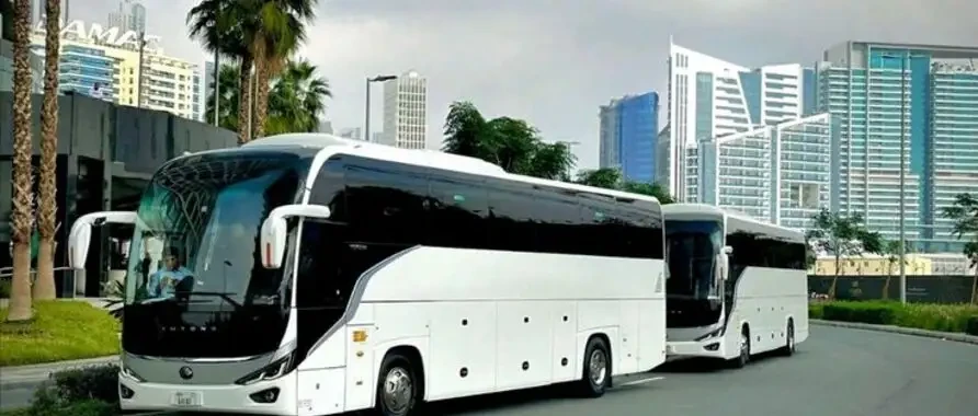 A white 16-50 seater bus parked in front of a building, available for rent in Dubai and Abu Dhabi.