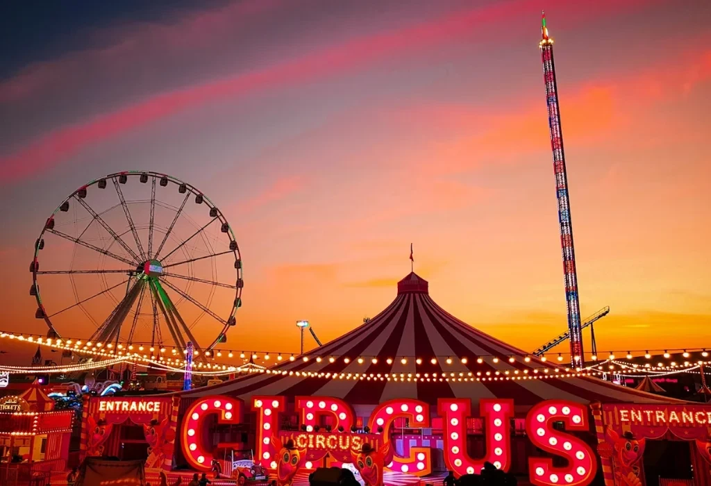 A red and white circus tent and a large Ferris wheel at Global Village Dubai during a pink and orange sunset.