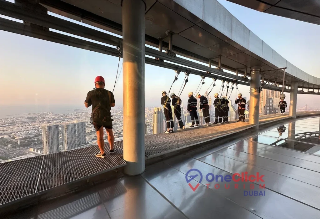 A group of tourists in safety gear standing on the open-air ledge of the Sky Views Observatory in Dubai during sunset.