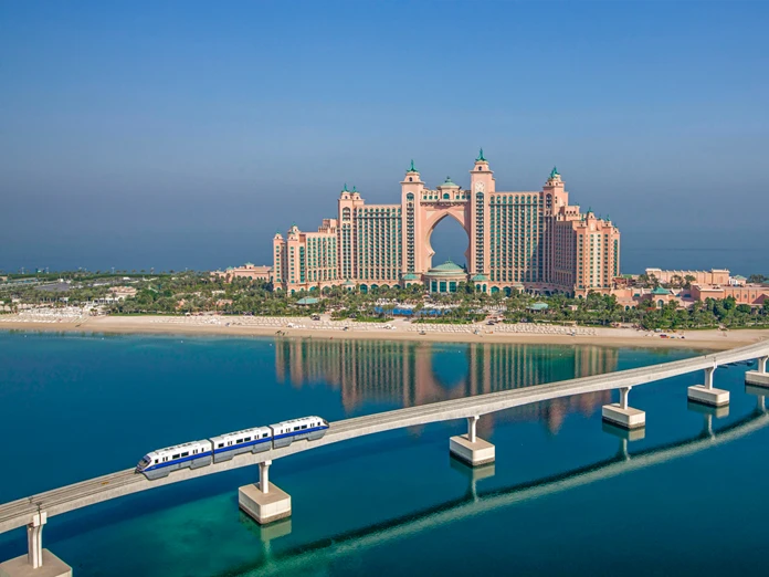 An aerial view of the Palm Jumeirah Monorail traveling along a concrete bridge over the blue ocean water with Atlantis, The Palm hotel in the distance.