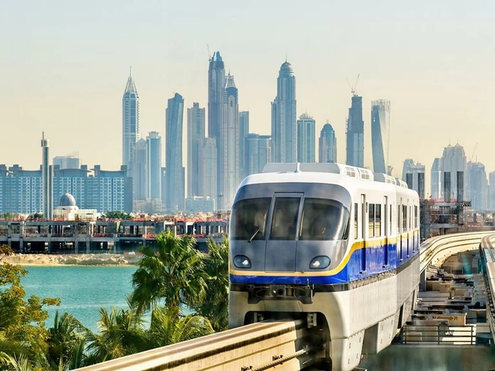 The Palm Jumeirah Monorail train traveling on an elevated track with the modern high-rise skyscrapers of the Dubai Marina skyline in the background.