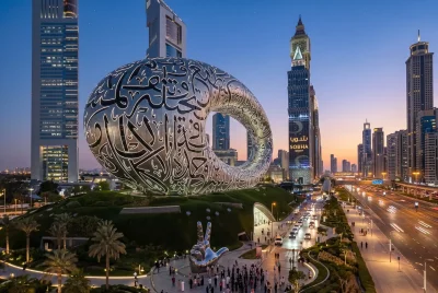 The Museum of the Future at night, showing its unique torus shape covered in glowing Arabic calligraphy, with a silver "Win, Victory, Love" hand gesture sculpture in the foreground.
