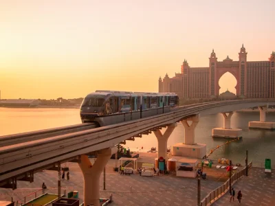 A side view of the Palm Jumeirah Monorail train on its track during a warm orange sunset with Atlantis, The Palm visible in the background.